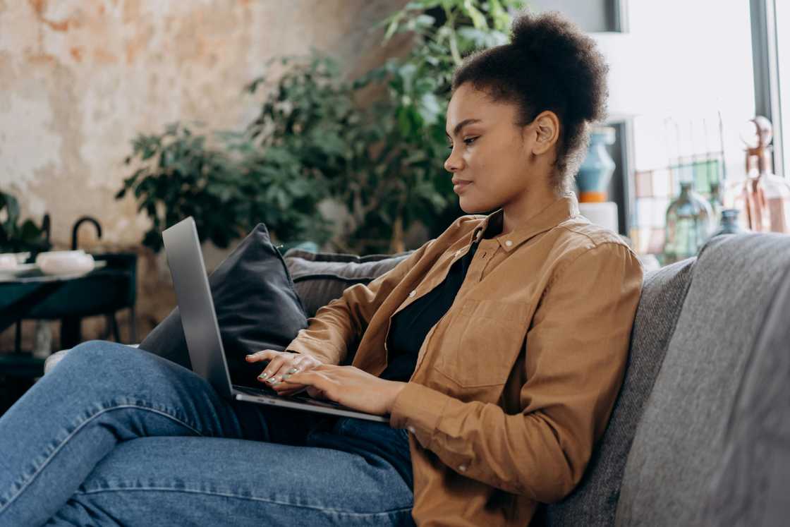 A woman in denim pants and a brown jacket is using a laptop on a grey sofa