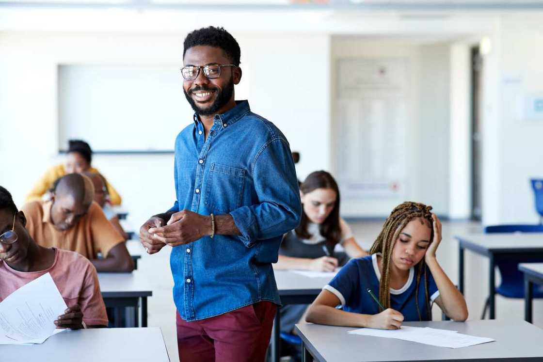 smiling young male supervisor standing amidst multi-ethnic students writing exam at desks in classroom